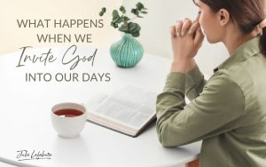 What Happens When We Invite God Into Our Days | woman sitting at desk praying with Bible open next to a cup of tea and a vase of flowers